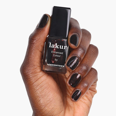 A person holds a bottle of LONDONTOWN Around the Fireplace nail polish in a deep, copper-flecked shade, with nails painted to match.