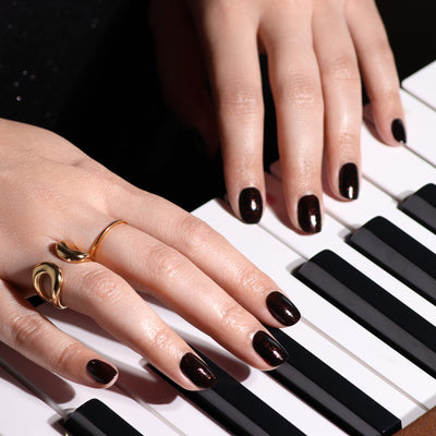 Close-up of two hands with deep, dark polished nails painted with LONDONTOWN's "Around the Fireplace" and adorned with gold rings, resting on a piano keyboard.