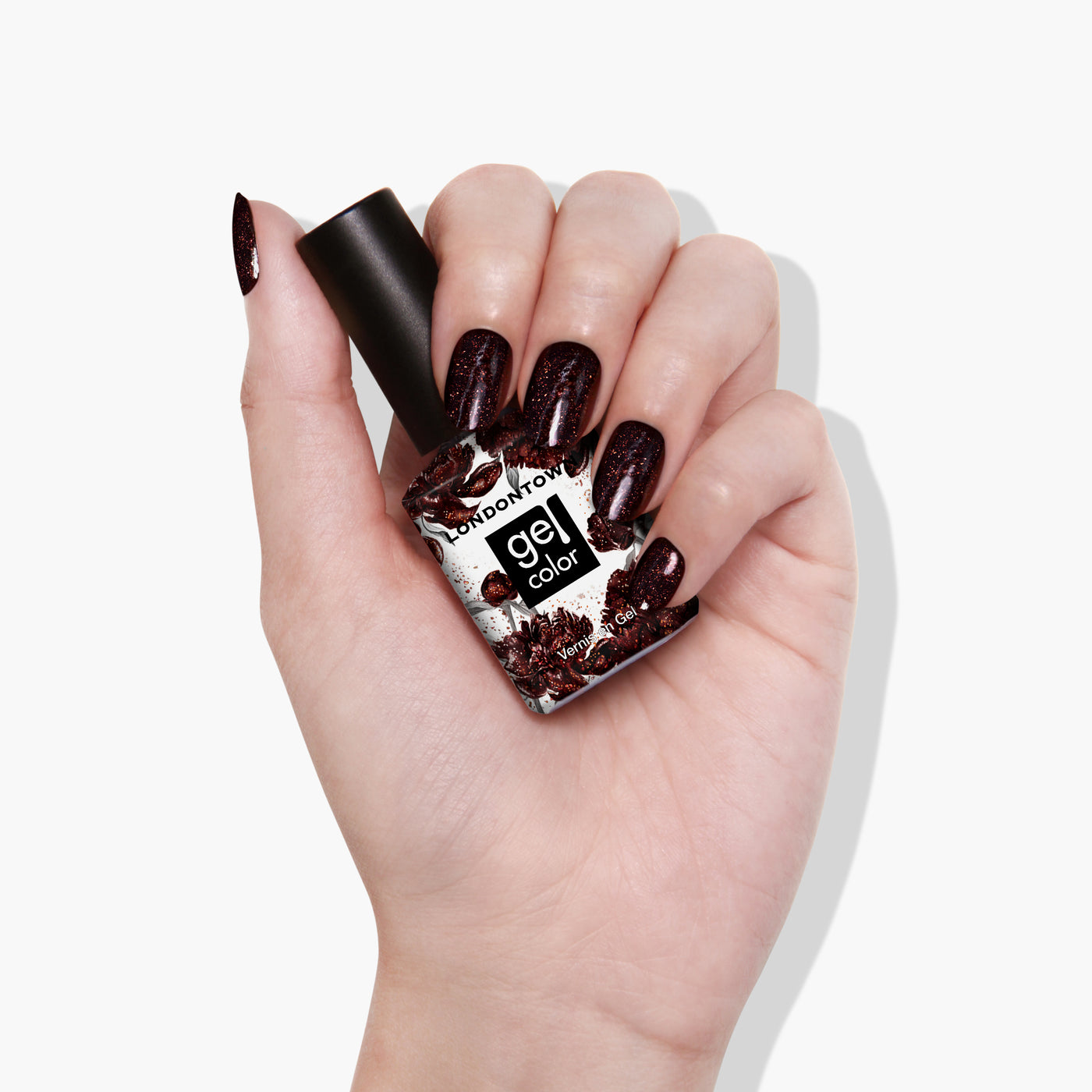 A hand with dark, glittery painted nails holds a bottle of LONDONTOWN's Around the Fireplace nail polish, boasting a deep, copper-flecked hue.