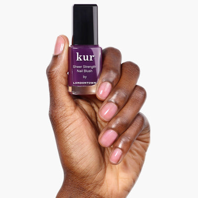 A hand holding a bottle of LONDONTOWN Sheer Strength Nail Blush – Concord with a purple label, showcasing nails painted in a light, sheer pink color. This 2-in-1 nail hardener also acts as a glow booster, giving your nails both strength and radiance.