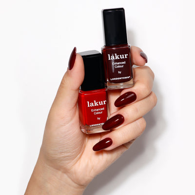 A hand with red painted nails holds two bottles from LONDONTOWN's Cozy Fireside Duo, featuring a vibrant red and a deep burgundy crème, perfect for stunning seasonal nails.