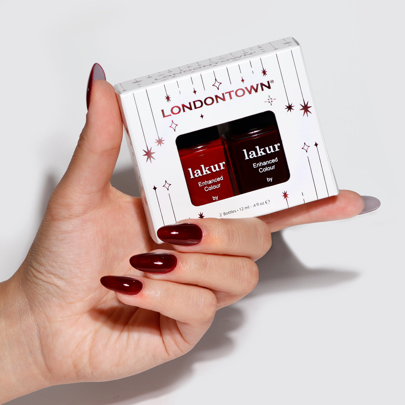 A close-up of a hand holding a package with two bottles from the Cozy Fireside Duo by LONDONTOWN, displaying rich deep red crèmes perfect for seasonal nails.