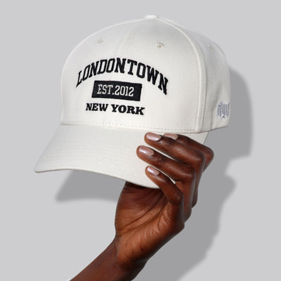 A hand holds a sun-smart, white Londontown Logo Adjustable Cap with "LONDONTOWN EST. 2012 NEW YORK" embroidered in black text on the front.