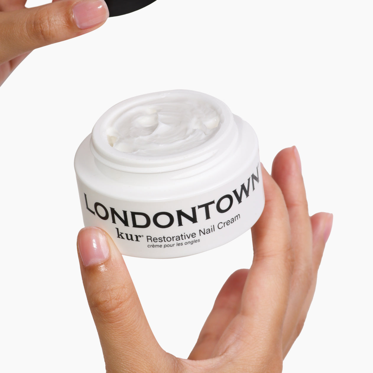 Hands holding an open jar of LONDONTOWN Restorative Nail Cream, a transformative moisturizer for dry nails, against a white background.