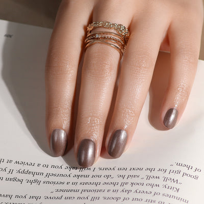 A hand adorned with LONDONTOWN's Nightcap metallic silver nail polish and multiple rings, including a delicate pearl ring, rests on an open book.