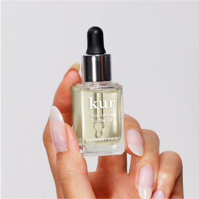 A hand holds a glass bottle of "Nourishing Cuticle Oil" by LONDONTOWN, featuring a black dropper cap. Infused with Florium Complex, this oil is crucial for nail restoration.