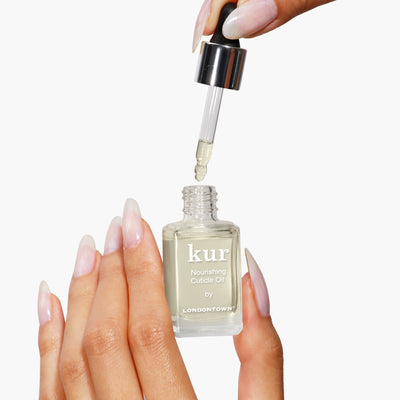 Hands gently using a dropper to apply LONDONTOWN's Nourishing Cuticle Oil, infused with the Florium Complex, from a small bottle for enhanced nail restoration.