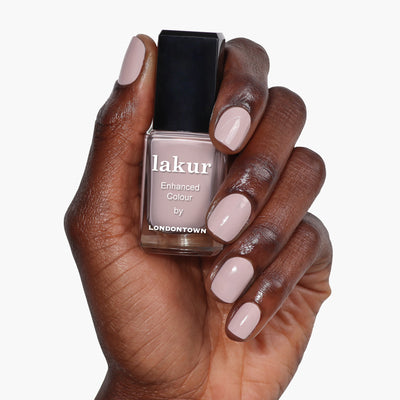 A hand with neatly manicured nails painted in a cool pastel beige color holds a bottle of "Off the Record" nail polish by LONDONTOWN in a matching shade.