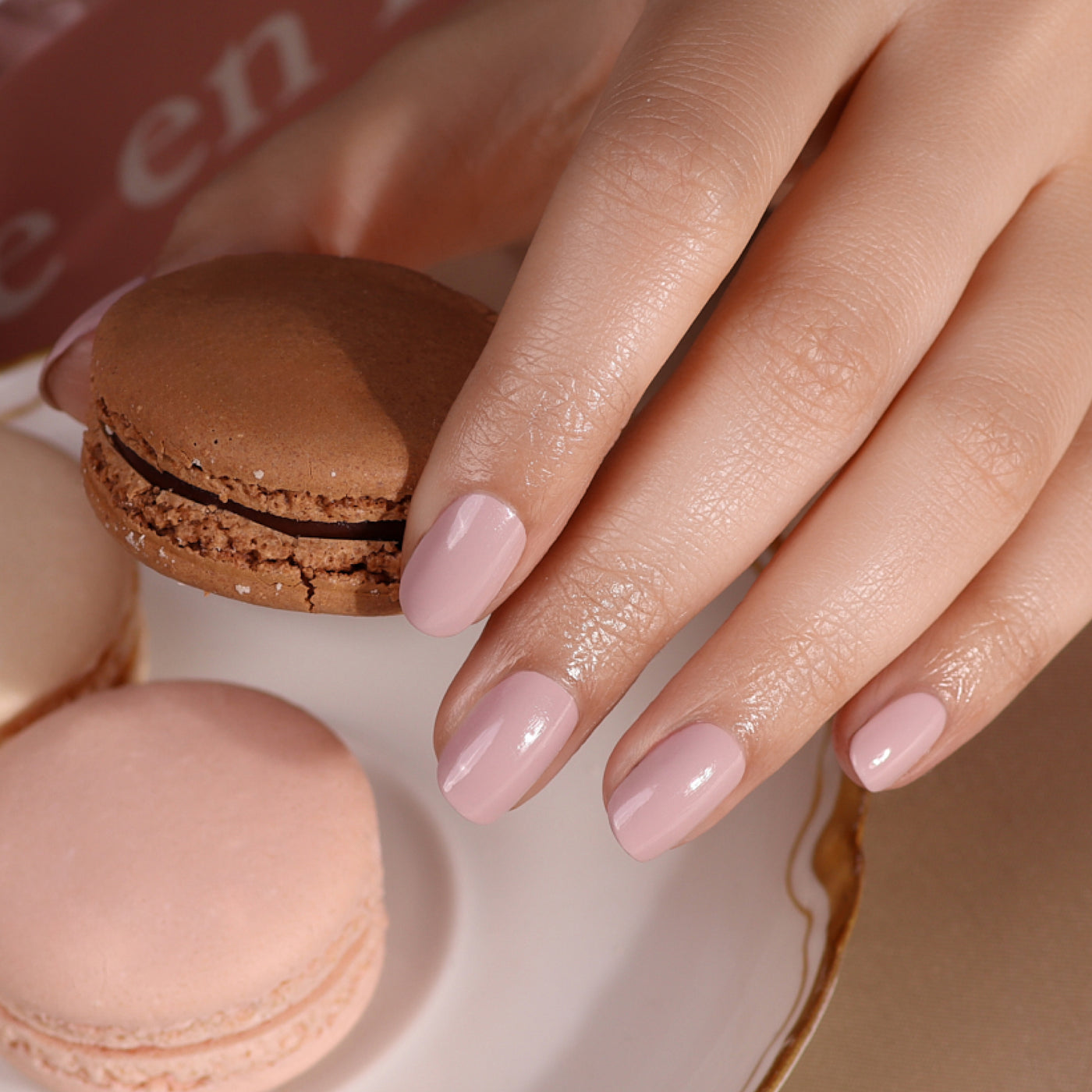 A hand adorned with LONDONTOWN's Off the Record light pink nail polish holds a brown macaron, while three other macarons—two pastel pink and one white—sit coolly on the plate below.