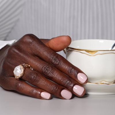 A close-up of a hand with chic, Off the Record mauve manicured nails from LONDONTOWN, adorned with a large ring, resting on a white surface next to a white and gold teacup with a spoon inside.