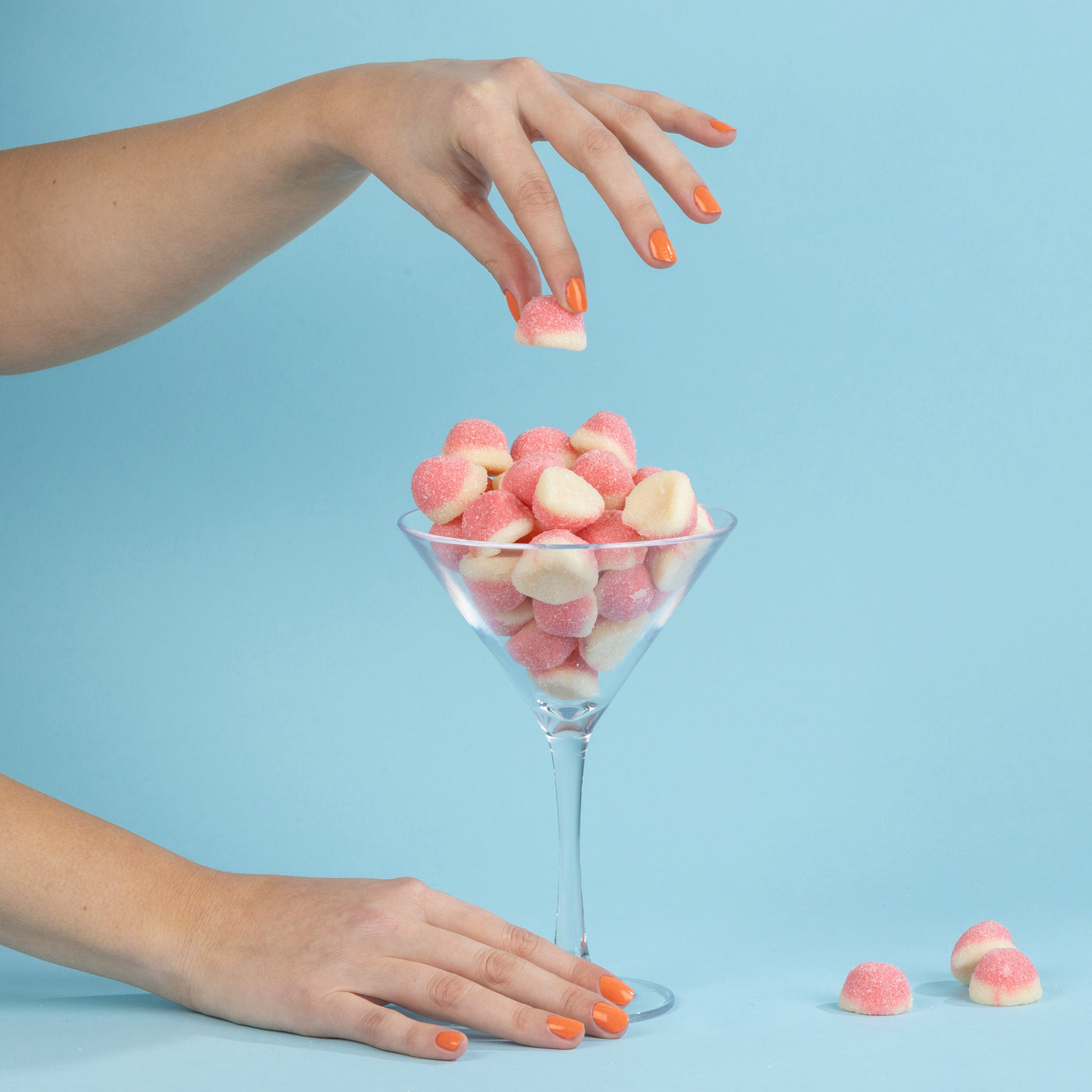 Two hands, adorned with the vibrant Paloma orange nail polish by LONDONTOWN, hold and grab pink sugar-coated candy marshmallows from a martini glass against a blue background.