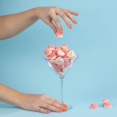 A hand with orange nails drops LONDONTOWN's Paloma, a pink and white gummy candy, into a martini glass brimming with similar treats, all set against a light blue background that captures the vibrant essence of California peach.