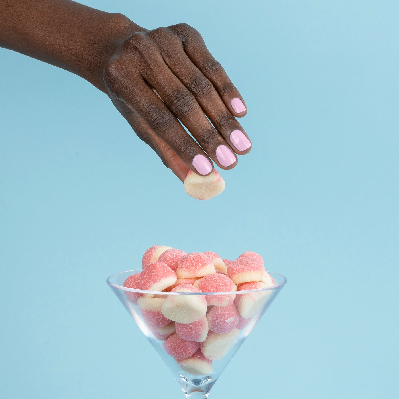 A hand adorned with LONDONTOWN's Pink Palm nail polish holds a gummy candy above a martini glass brimming with more gummy candies, set against a light blue background.