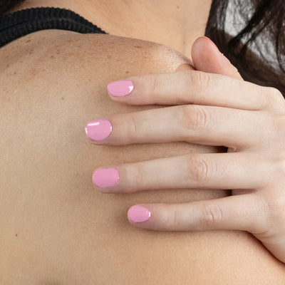 A close-up of a person's hand adorned with LONDONTOWN's Pink Palm nail polish, a delicate pastel pink reminiscent of a carnation, resting gently on their shoulder. The person is wearing a sleeveless dark top.