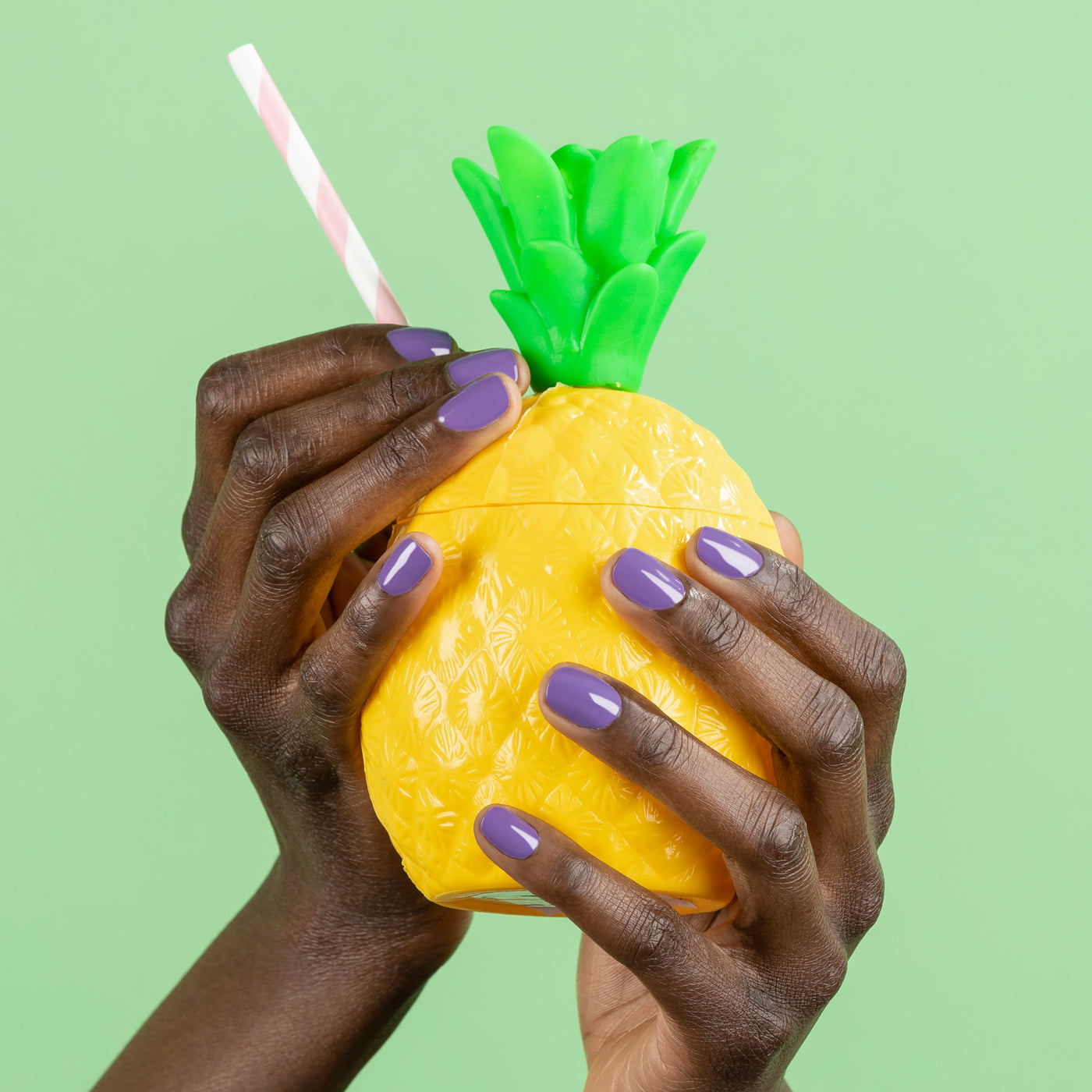 Close-up of hands featuring Road Trip lavender nail polish by LONDONTOWN, holding a yellow pineapple-shaped drink container with a green top and a psychedelic striped straw, set against a green background.