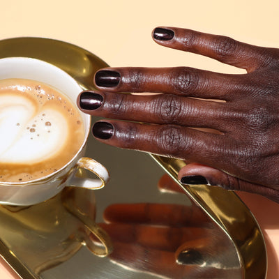 A hand adorned with LONDONTOWN's Spilled Wine nail polish rests beside a cup of coffee on a gold-colored tray, basking in the soft glow of candlelight.