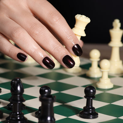 A hand with Spilled Wine-painted nails by LONDONTOWN moves a white rook on a green and white chessboard, illuminated by candlelight. Black chess pieces are positioned in the foreground.