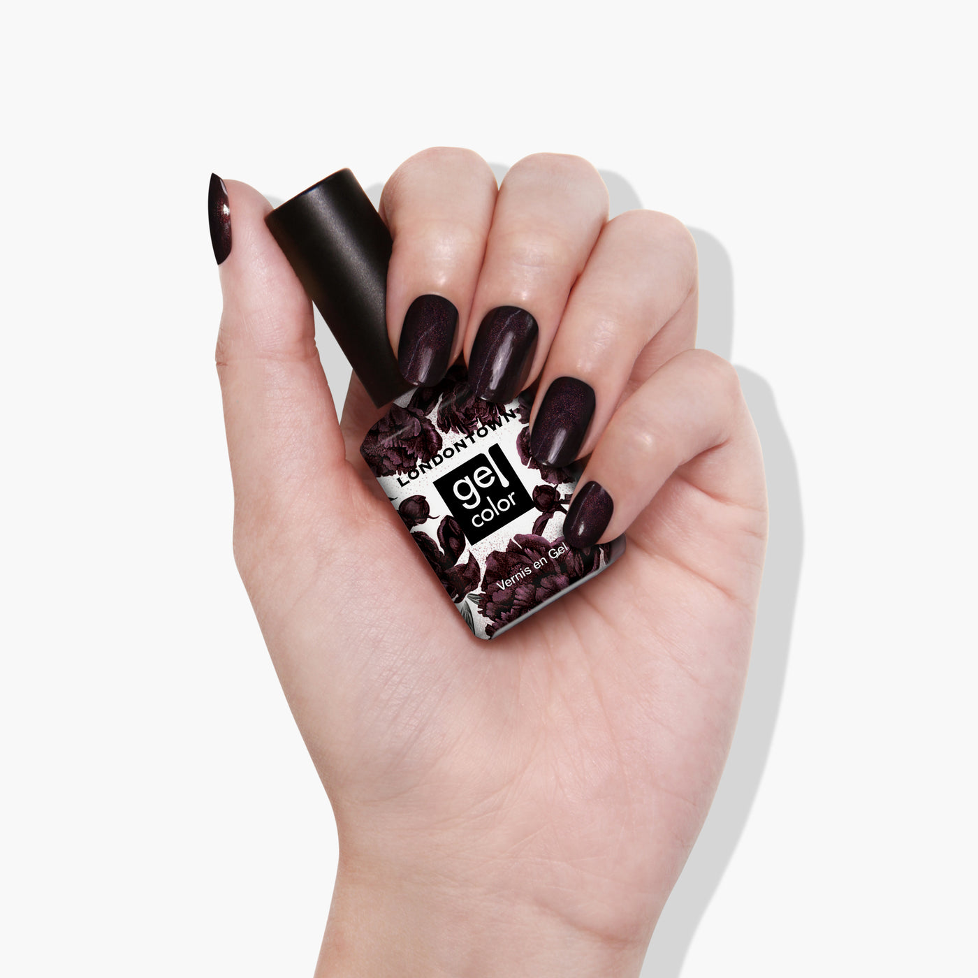 A hand with dark purple nail polish is holding a bottle of LONDONTOWN Spilled Wine Gel Color nail polish, featuring a floral design on the label that evokes the richness of a deep pinot noir.