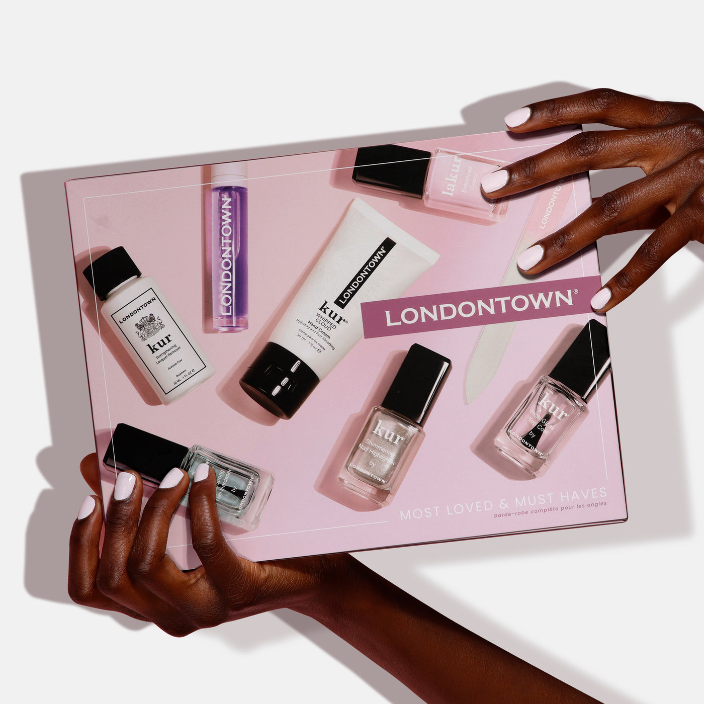 Hands hold a LONDONTOWN Most-Loved & Must-Haves Set showcasing nail polish, remover, and hand cream against a pink background—ideal for holiday season pampering.