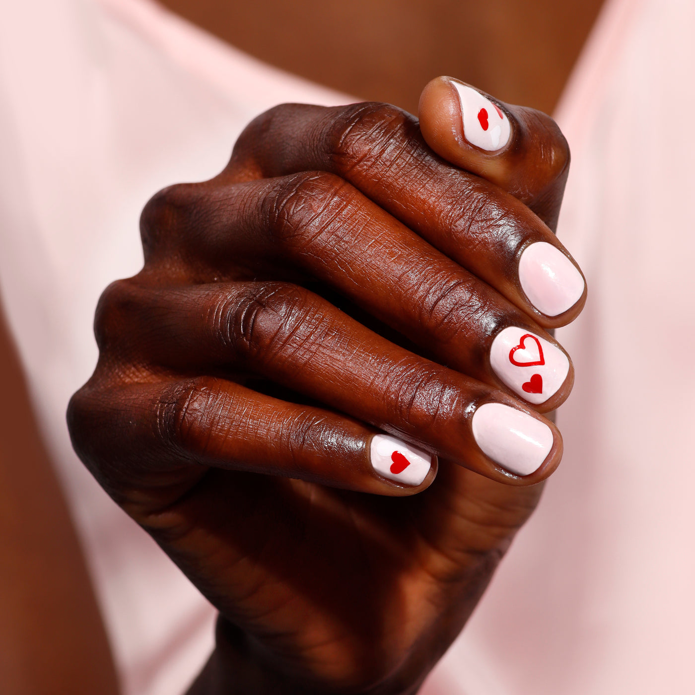 A hand showcases a mani with LONDONTOWN's Love & lakur Valentine’s Day Set, featuring pink polish adorned with red heart designs on a soft pink background.