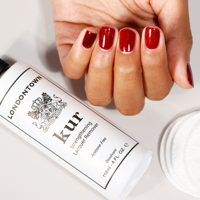 A hand with red-painted nails holds LONDONTOWN's Strengthening Lacquer Remover, along with cotton pads. This vegan and cruelty-free nail care essential ensures gentle and effective cleaning while serving as an acetone-free remover.