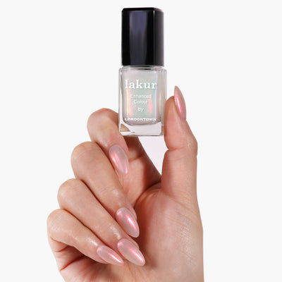 A hand with long, manicured nails painted in LONDONTOWN's Aurora Chrome, a shimmering pink crème-to-chrome shade, holds a lakur nail polish bottle. The sleek lacquer finish complements the elegant look against a plain white background.