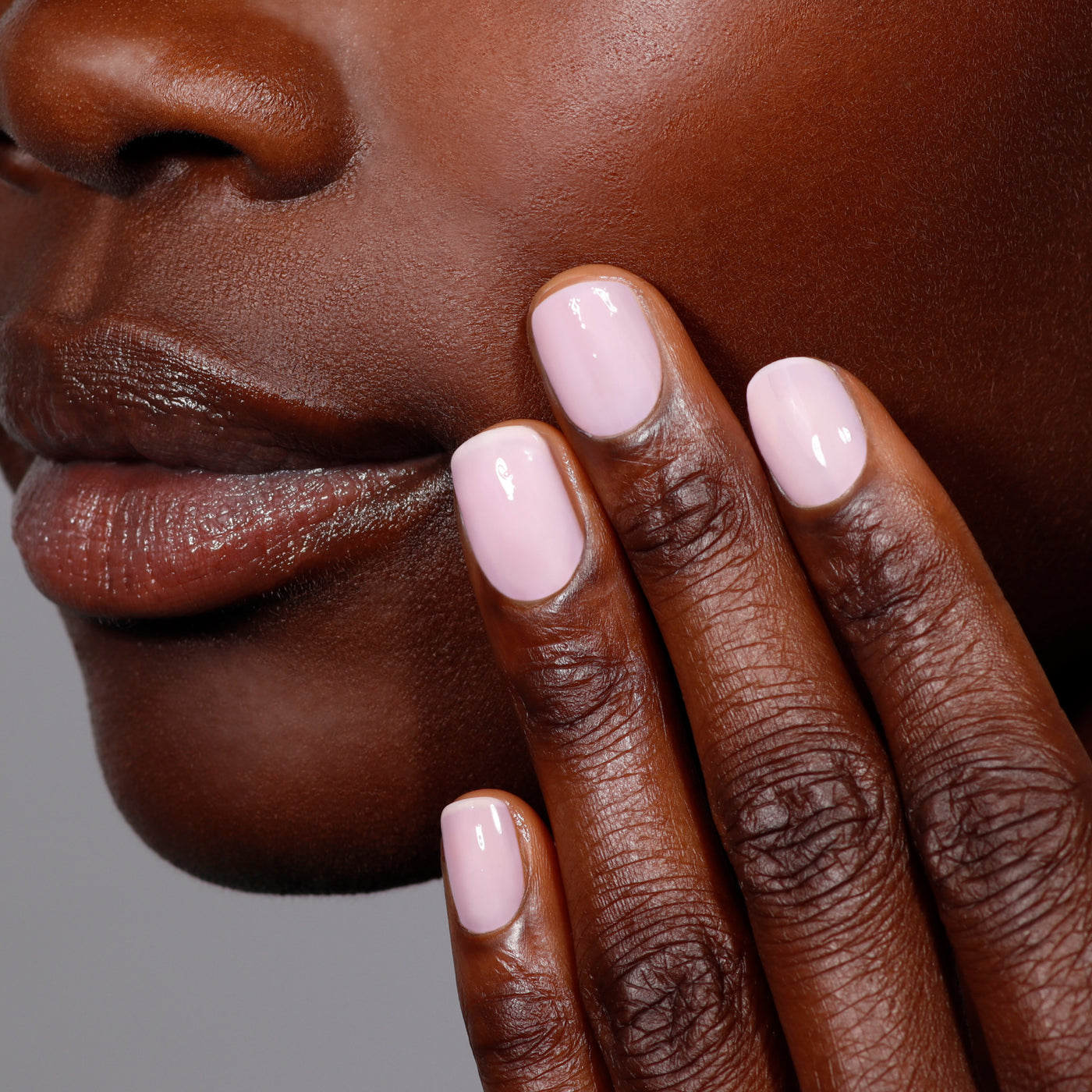 A close-up image features a person with versatile Magnolia Haze nails in petal pink by LONDONTOWN, gently touching their cheek. Their lips and chin are also visible, highlighting a smooth complexion.