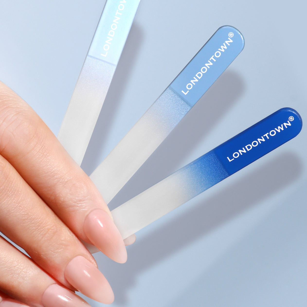 A hand displays a Mini Nail File Set from LONDONTOWN, featuring three glass files with blue gradient handles, against a light blue background, highlighting how they perfectly prep your nails.