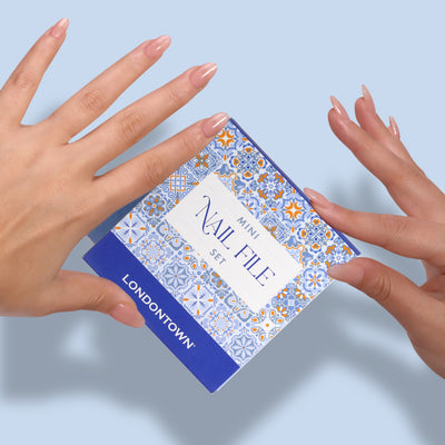 Two manicured hands hold a LONDONTOWN Mini Nail File Set box against a light blue background. The box, decorated with a blue and orange tile pattern, is filed up and ready to go.