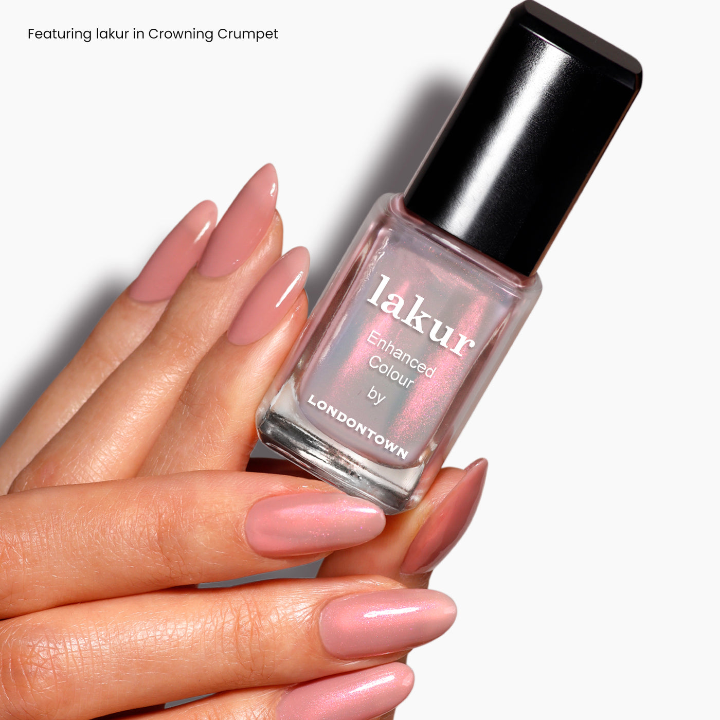 A hand with glossy, long, almond-shaped nails painted in a soft pink shade holds a bottle of LONDONTOWN nail polish labeled "Aurora Chrome," showcasing its luxe lakur finish.