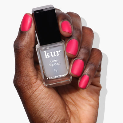 A hand with pink nails holds a bottle of LONDONTOWN Matte Top Coat (Professional), offering an ultra-luxe, suede-like finish.