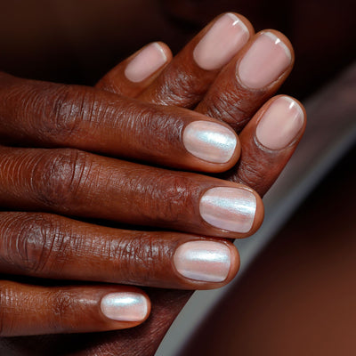 Close-up of hands with dark skin and manicured nails showcasing LONDONTOWN's Moonstone Chrome finish. Nails are painted glossy light pink, with some having a crème-to-chrome metallic effect.