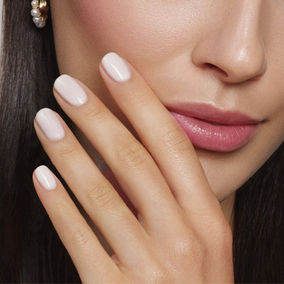 A person with long brown hair and pearl earrings gently touches their face, showcasing light pink nails polished with "Princess Awaits" by LONDONTOWN. This vegan, cruelty-free nail polish features a sheer white shimmer, ensuring beauty without compromise.