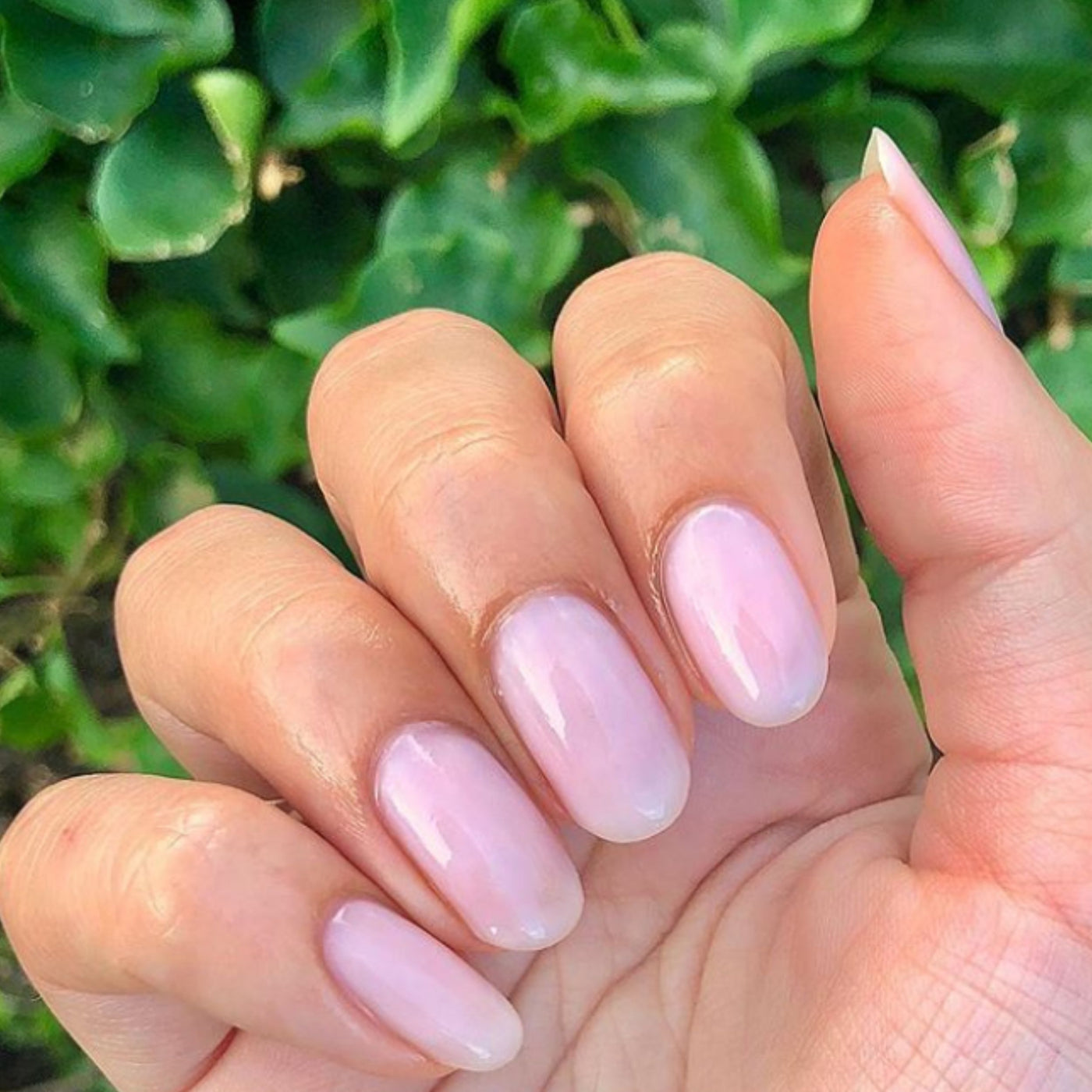 A hand with nails polished in LONDONTOWN's Invisible Crown, a soft sheer pink, is held against lush green leaves, subtly dazzling the serene scene.