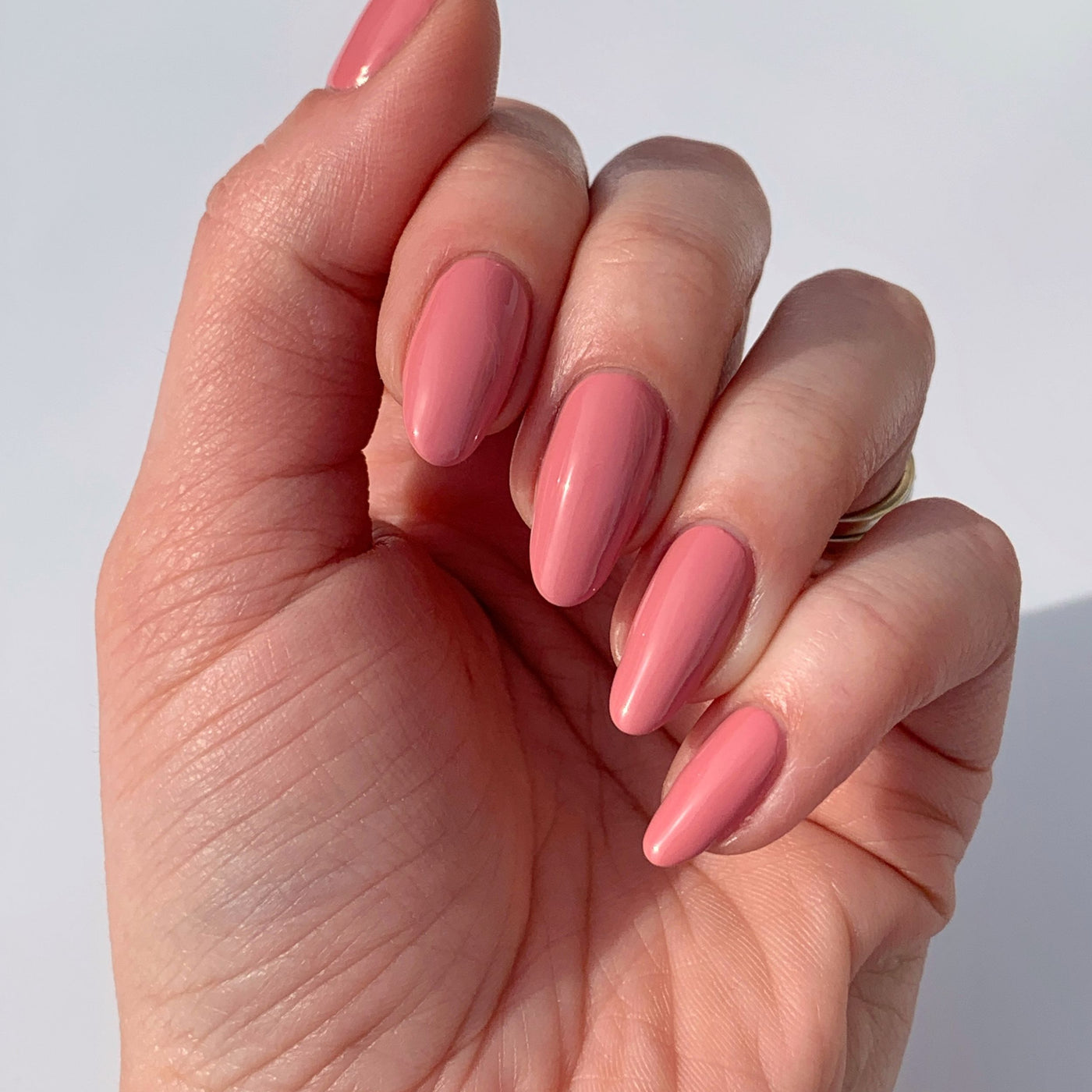 A hand with long almond-shaped nails painted in LONDONTOWN's Mojave Mauve, a glossy pink vegan polish, is displayed against a plain background, showcasing an elegant cruelty-free manicure.