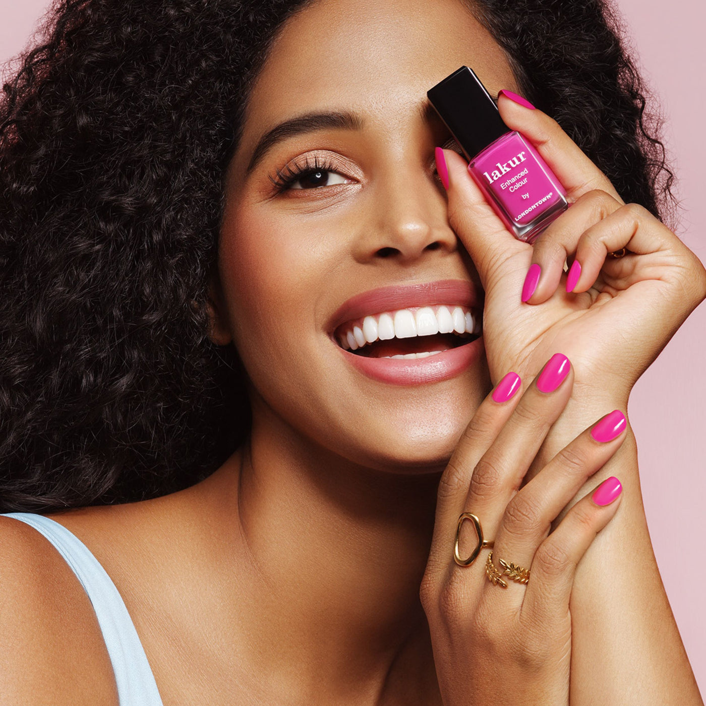 A curly-haired person smiles while holding a bottle of LONDONTOWN's Summer Fling nail polish in a shocking pink shade near their face. The vegan, cruelty-free nails complement their light top perfectly.