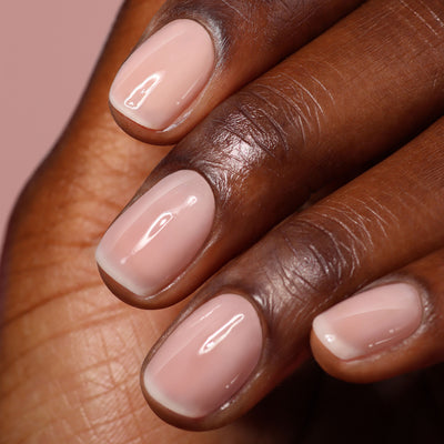 A close-up of a hand with a light pink gel-like mani on short, neatly groomed nails, finished with LONDONTOWN's Gel Genius Top Coat against a soft pink background.