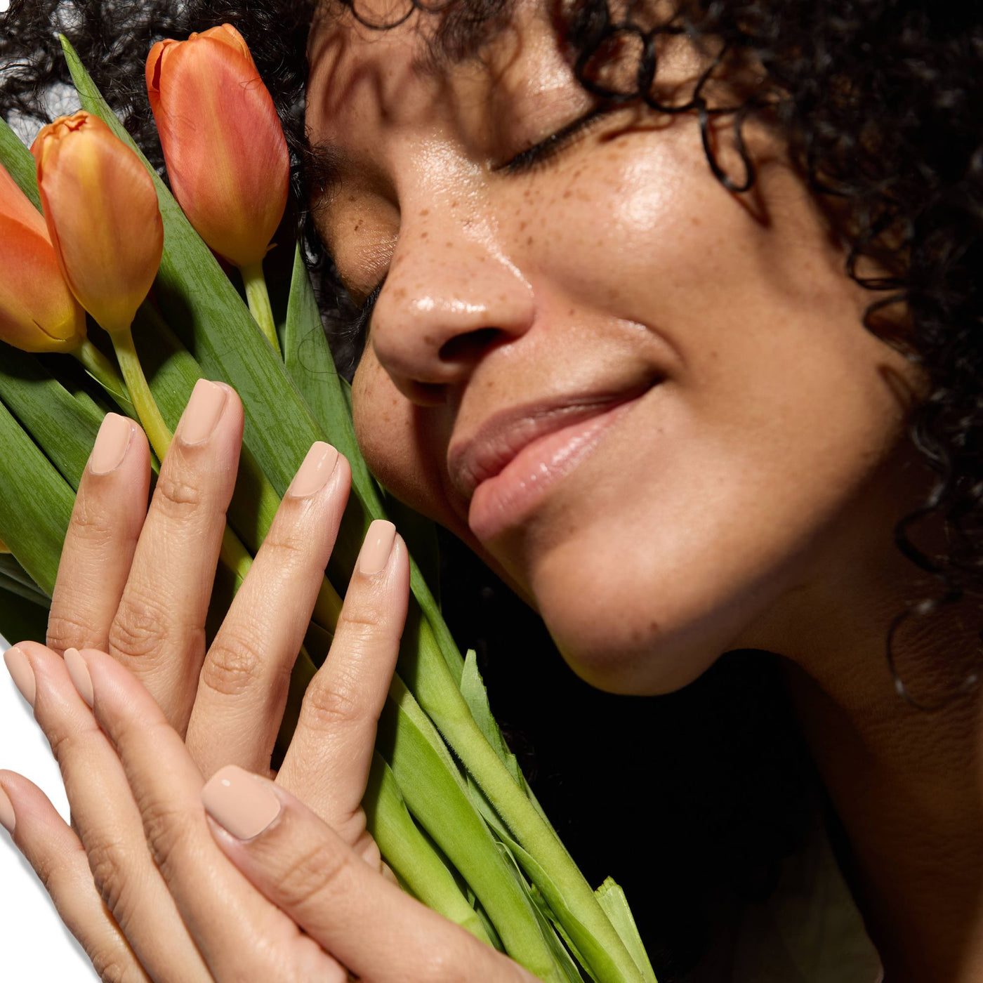 A person with curly hair smiles gently while holding three orange tulips, their face reflecting the shine of the LONDONTOWN Protective Top Coat (Professional) petals.