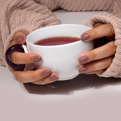 Wearing a beige sweater, a person holds a white cup of tea with both hands, savoring the moment like an afternoon break finished with LONDONTOWN's Flash Dry Top Coat.