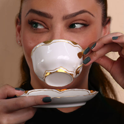 A person with a Gel Genius Top Coat manicure by LONDONTOWN holds a white and gold teacup and saucer near their mouth, gazing to the side.