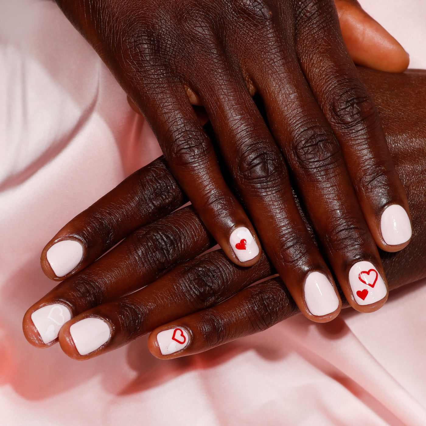 Hands with LONDONTOWN's Invisible Crown white nail polish and red heart designs rest on silky, sheer pink fabric.