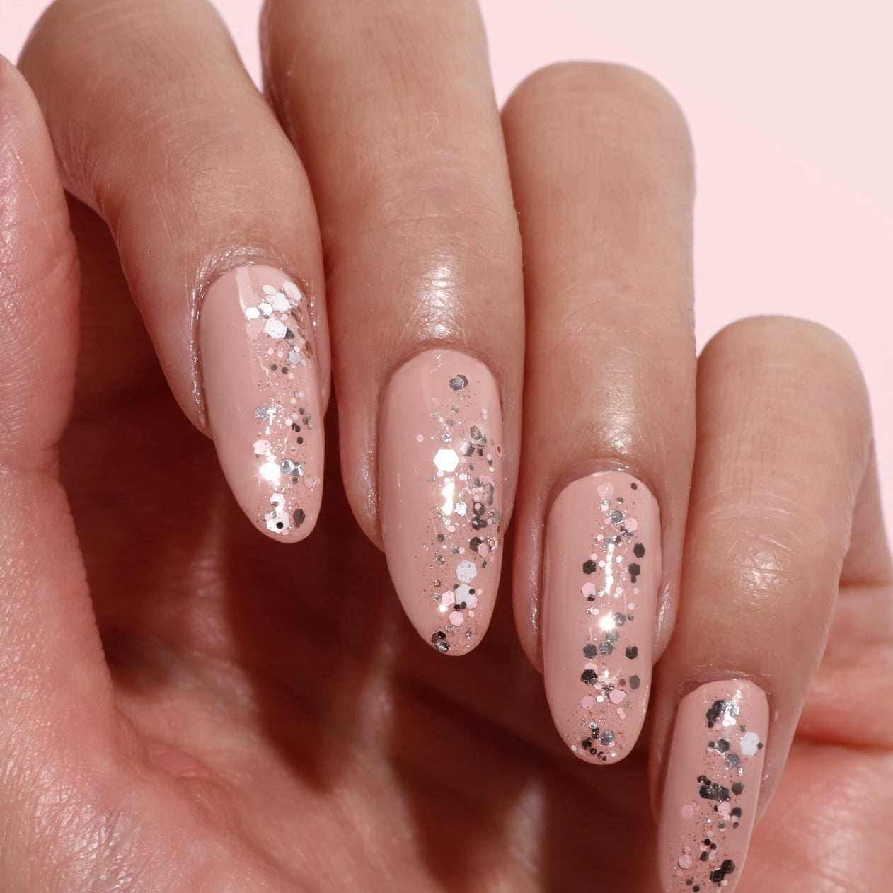 A hand showcases nude nails enhanced with LONDONTOWN's Snowglobe Confetti Topper, adding a sparkling silver glitter finish, set against a light background.