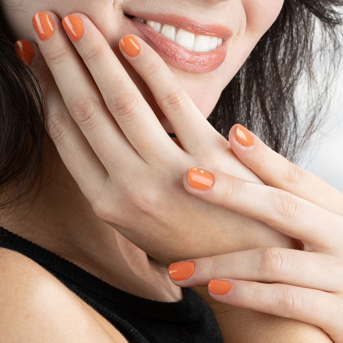 A person with orange nail polish touches their face, smiling and highlighting the high shine and durability of LONDONTOWN's Protective Top Coat (Professional).