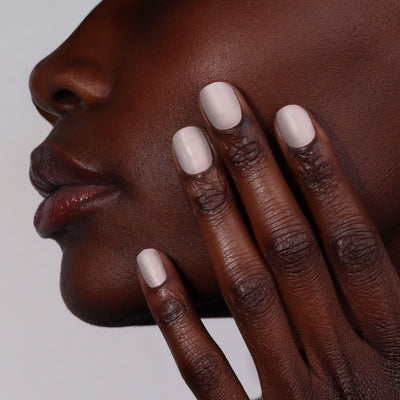 Close-up of a person touching their face with fingers, each nail painted with Beaumont (Professional) by LONDONTOWN greige polish, against a plain background.