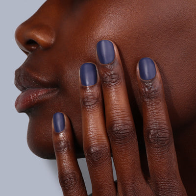 A person's hand, adorned with LONDONTOWN's Matte Top Coat (Professional) in an ultra-luxe, suede-like blue finish, rests against their face, showcasing smooth skin and impeccably groomed nails.