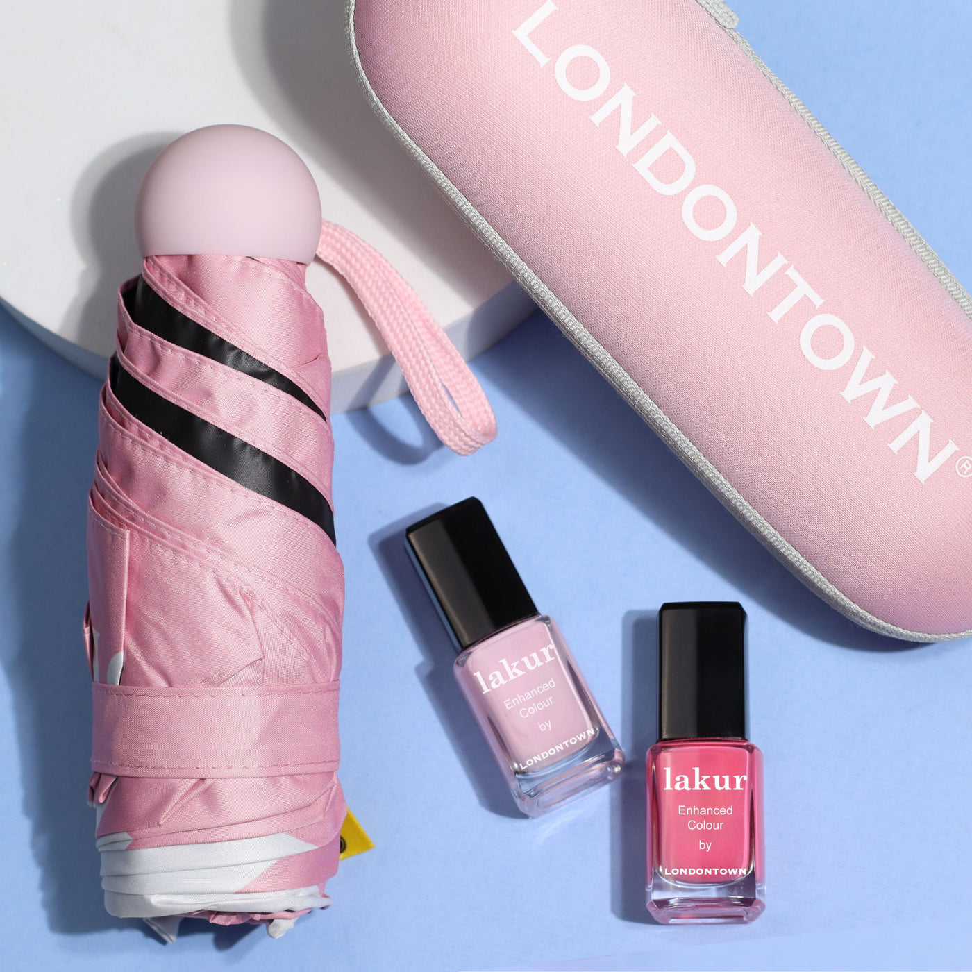 A folded Signature Mini Umbrella in pink, a pink LONDONTOWN case, and two bottles of lakur nail polish—one in light pink and the other in darker pink—are arranged on a blue surface. Perfect as your travel companion, this portable set ensures you're always prepared with your favorite essentials.