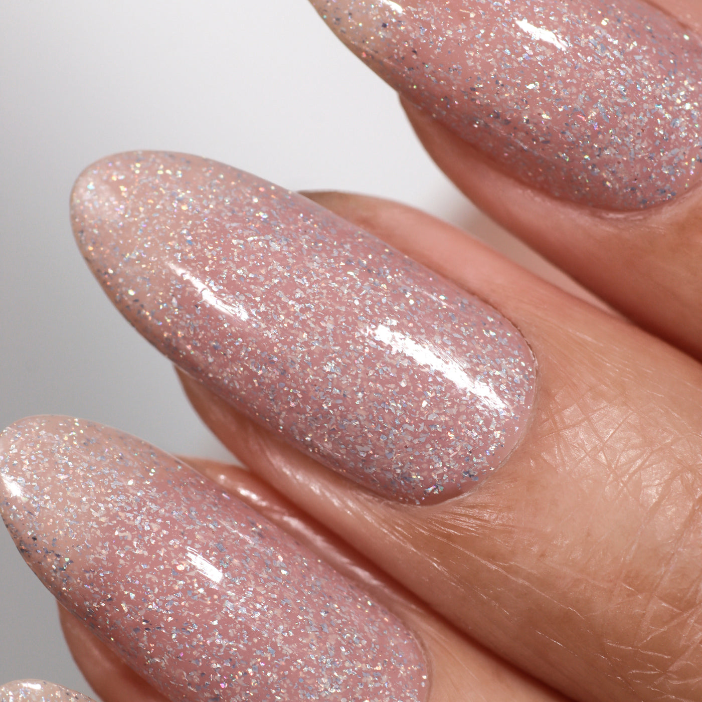 Close-up of fingernails with a glossy manicure using LONDONTOWN's Champagne in Bed, showcasing a neutral pink shade with silver glitter and holographic sparkle.