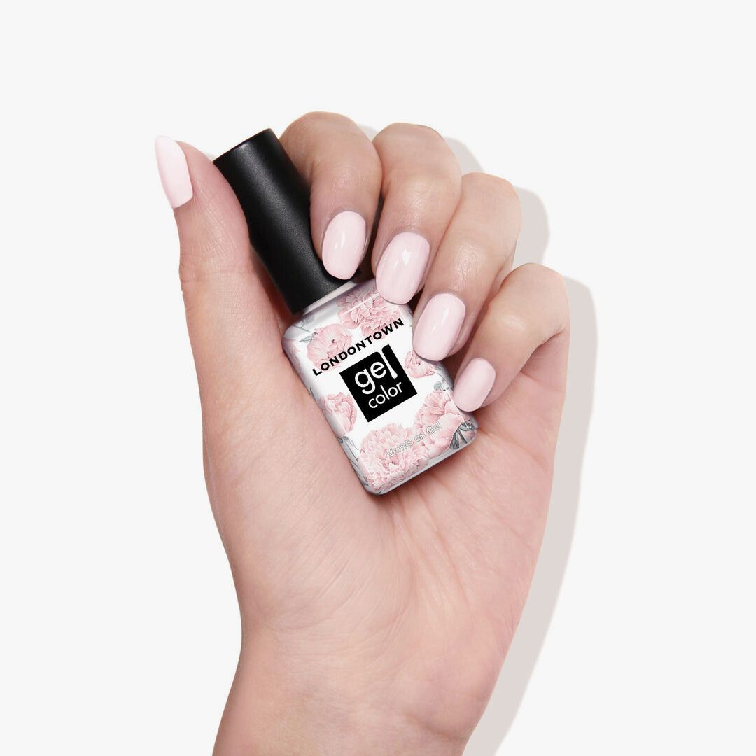 A hand displays the LONDONTOWN Macaroon at Noon gel polish bottle with pastel powder pink nails against a white background, highlighting this subtle hue's delicate charm.