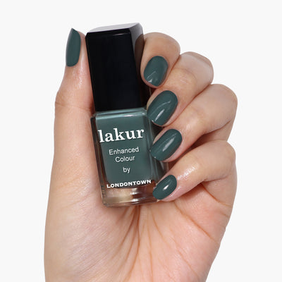 A hand holding a bottle of LONDONTOWN Velvet Drapes nail polish in a sumptuous olive green color, with matching painted fingernails.