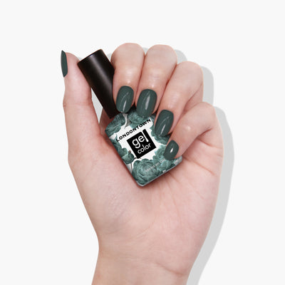 A hand with manicured nails is holding a bottle of LONDONTOWN Velvet Drapes nail polish in a sumptuous olive green shade.